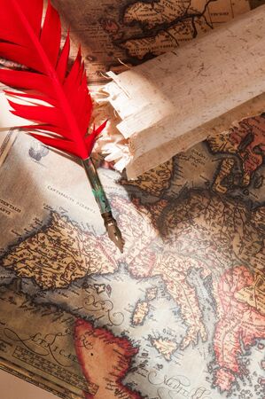 Red quill pen, a green inkwell and a rolled papyrus sheet with some old maps on a wooden tableの写真素材