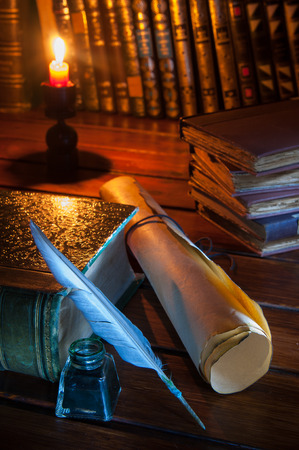 Quill pen and a rolled papyrus sheet on a wooden table with old booksの写真素材