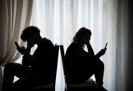 Silhouette of two teenagers with cell phones sitting and facing away from each otherの写真素材