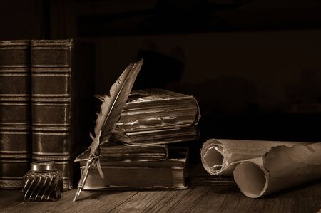 Quill pen and a rolled papyrus sheet on a wooden table with old booksの写真素材