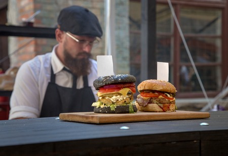 KIEV, UKRAINE - 16 AUG 2015: Festival street food. Two hamburger stand on the board with tags. In the background, cook with a beard, wearing glasses and a cap.のeditorial素材