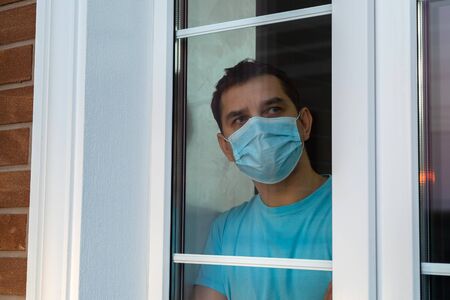 Isolation at home for self quarantine. Young man in medical mask is looking out window. Guy stay home because of quarantine to prevent spreading of coronavirus. Coronavirus pandemic. Social distancingの写真素材