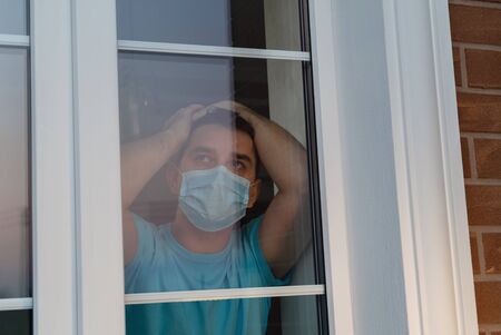 Man in a medical mask near window. Man tired of quarantine of Coronavirus. Sick sad man in medical protective face mask looking through window. Coronavirus pandemic covid-19 and quarantine concept.の写真素材