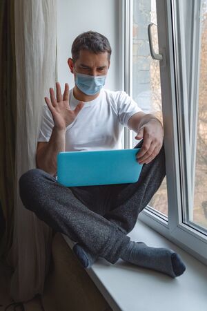 Young man in a blue protective medical mask sits on a windowsill with a laptopの写真素材