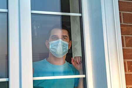 Infected man on self-isolation looks at street through the window of a house. Young brunette white european man at home on quarantine with medical mask near window white backround. Social distancing.の写真素材