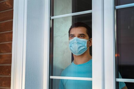 A man with a protective medical mask at the window. Quarantined, isolated from society. Young attractive man in a blue clothes with medical mask showing a nameplate with the lettering Stay at home.の写真素材