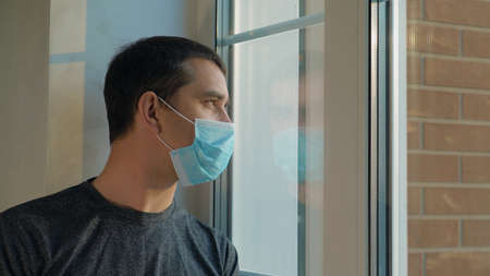 Quarantine self-isolation. Sad young man in a medical mask who looks out the window through the window. Infected man in medical mask on self-isolation looks at the street through the window of a houseの写真素材