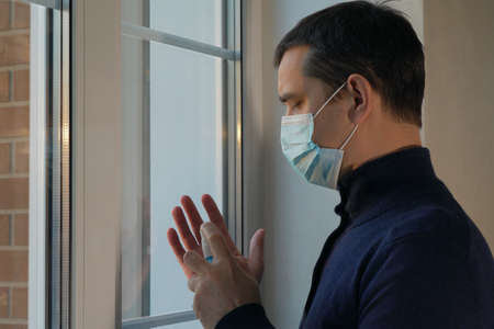 Quarantine self-isolation. Sad young man in a medical mask who looks out the window through the window. Infected man in medical mask on self-isolation looks at the street through the window of a houseの写真素材