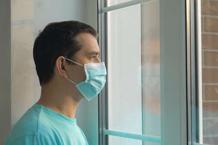 Quarantine self-isolation. Sad young man in a medical mask who looks out the window through the window. Infected man in medical mask on self-isolation looks at the street through the window of a houseの写真素材
