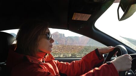 Young woman is driving a car on a sunny day, outside the window is a mountain landscape the Red Rock Canyon. Woman in a red jacket driving, close up. Woman with a serious look driving a car.の写真素材