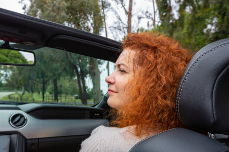 Woman with red curly hair sits in a convertible, rear view, CLOSEUP. Woman Passenger On Road Trip In Convertible Car. Portrait of smiling young woman in convertible car. Travel and holidays conceptの写真素材