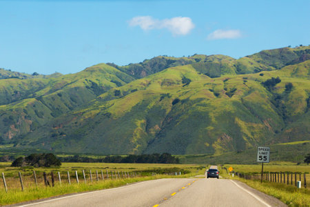 Road stretches into the distance against the backdrop of green meadows and mountains. Asphalt road with a dividing strip goes into the distance. Car drive far along the highway, rear viewの写真素材