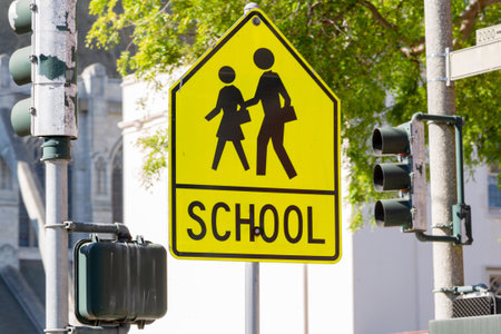 Yellow warning road sign for a school zone at a road intersection with a street traffic light in the background. Yellow School Zone Street Sign, Close-up. School Road Sign with Children, Foregroundの写真素材