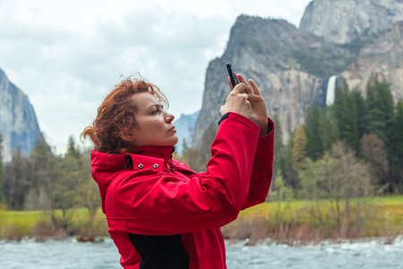 Young woman takes pictures of a natural landscape in Yosemite Park, California with a smartphone camera. Yosemite Falls in Springtime. Scenic view of rocky mountains against skyの写真素材