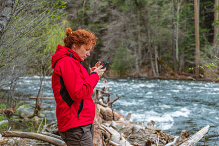 Young white caucasian woman checks her mobile phone in nature while traveling. Beautiful young woman holding mobile phone walking near river in forest. Phone distracting from travel, concept.の写真素材