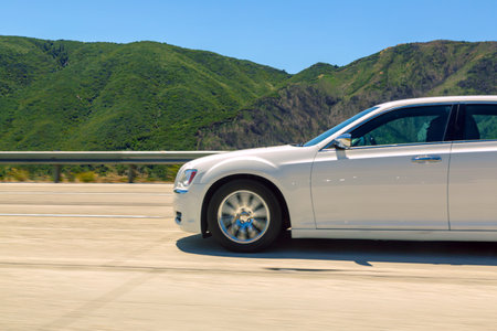 White car driving fast on mountain highway. Car moving at speed on asphalt mountain road. Motion and journey experience. Mountainous landscape visible in background. Road trip and travel themesの写真素材