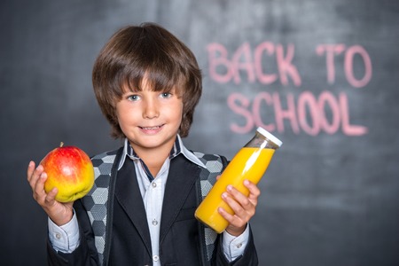 Close-up of little school boy with apple and juice standing near blackboard with words \"back to school\" on itの写真素材