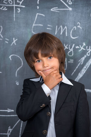 Cute little school boy with messy hair solving difficult equations written on blackboardの写真素材