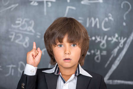 Cute little school boy with messy hair solving difficult equations written on blackboardの写真素材