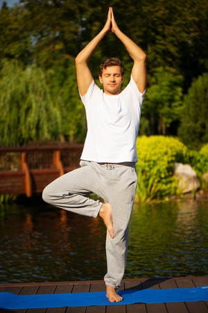 Young man practising yoga and standing in asana with a beautiful green scenery of park on backgroundの写真素材