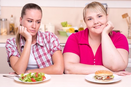 Two contradictions. Young woman with well set up figure and young overweight woman eating healthy and junk foodの写真素材