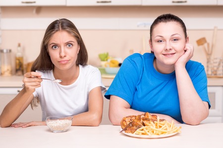 Two contradictions. Young woman with well set up figure and young overweight woman eating healthy and junk foodの写真素材
