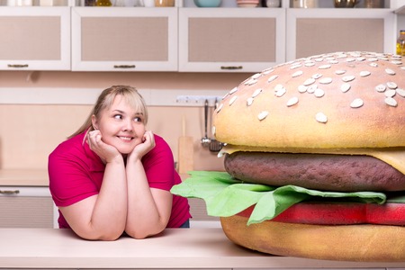 Young smiling overweight woman standing in kitchen and looking at huge hamburgerの写真素材