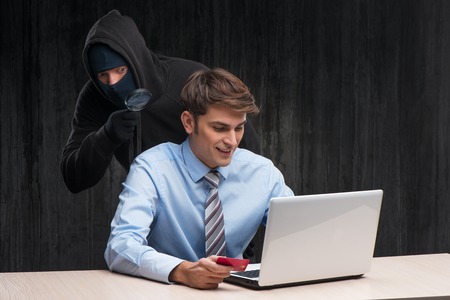 Masked man using hand-glass to shop online with stolen card. Internet theft conceptの写真素材