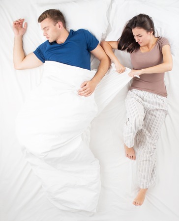 Young couple sleeping in big white bed. Top view photo. Man and woman can not divide a blanket. Angry woman trying to take a blanketの写真素材