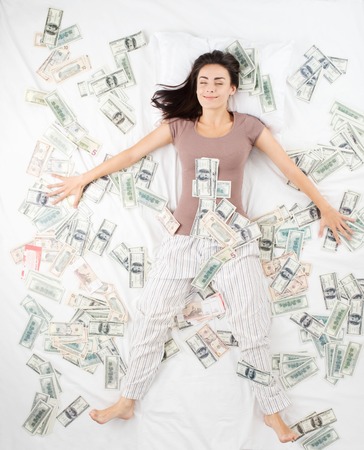 Happy smiling young woman sleeping in a bed full of banknotes. Concept for dreams of winning jackpot in lotteryの写真素材