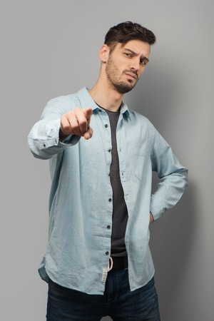 Thoughtful young man pointing at camera, standing on grey backgroundの写真素材