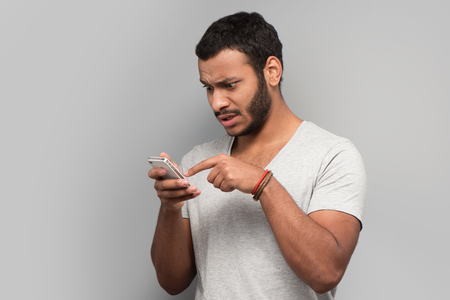 Young mixed race man trying to use smart phone, standing on grey backgroundの写真素材