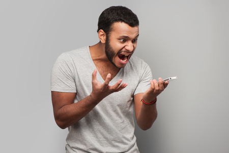 Young mixed race man screaming at phone, standing on grey backgroundの写真素材
