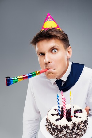 Funny close up picture of young office manager with party hat and blower. Man holding cake and looking at cameraの写真素材