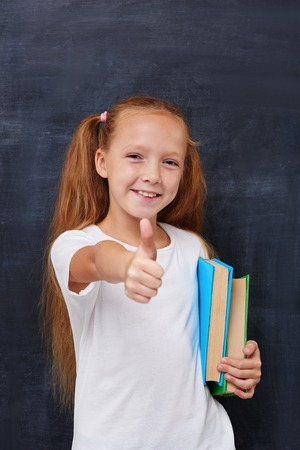 Beautiful redhead girl thirsted for knowlege looking at the camera, smiling and showing thumbs up against the blacboard background,expressing positivity, school educational conceptの写真素材