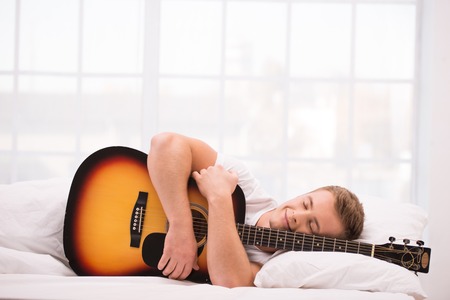 Photo of handsome young man. He is comfortably curled under a white blanket and holding guitarの写真素材