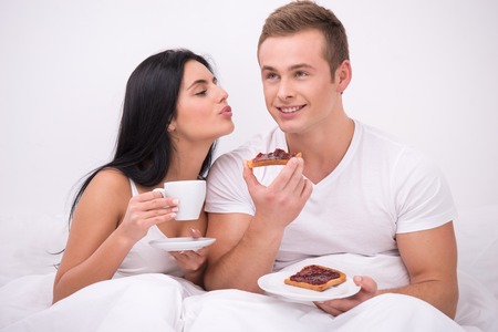 Young couple sitting under white blanket early in the morning and having their breakfast in bed. Woman with cup of coffee kissing her husband while he eating toasts with jamの写真素材