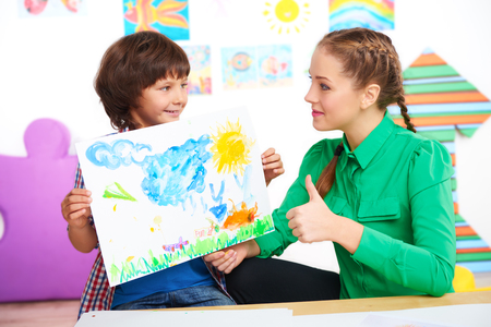 Young teacher looking at finished drawing of boy and showing thumb up. Lesson in preschool center for childrenの写真素材