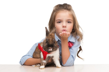 Little beautiful girl blowing a kiss to camera and holding brown bunny with red bow tieの写真素材