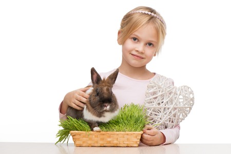 Beautiful little girl sitting isolated on white background with cute brown bunny in grass and holding white heartの写真素材
