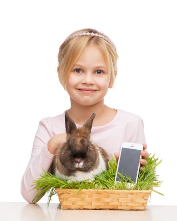Beautiful little girl sitting isolated on white background with cute brown bunny in grass and holding telephoneの写真素材