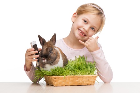 Little girl sitting isolated on white background with cute brown bunny in grass and showing call me signの写真素材