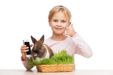 Cute girl sitting isolated on white background with brown bunny in grass and showing call me signの写真素材