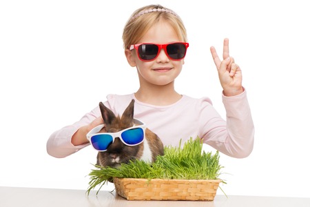 Funny picture of little smiling girl showing victory sign and brown bunny in sunglasses, isolated on whiteの写真素材