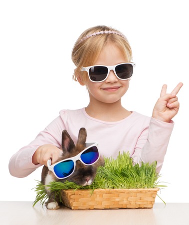 Photo of little smiling girl showing victory sign and brown bunny in sunglasses, isolated on whiteの写真素材
