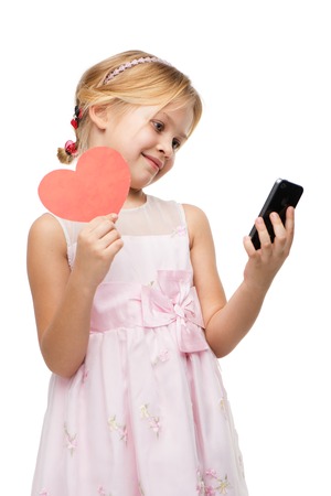 Beautiful little girl wearing pink dress, looking at the phone. Isolated on white background. Holding red paper heartの写真素材