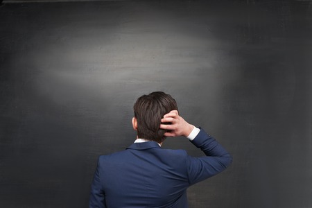 Photo of young pensive businessman on blank chalkboard background. Man standing with his back to the cameraの写真素材