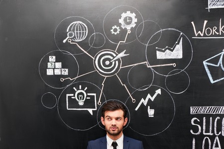 Young businessman standing on dark chalkboard background. Colorful symbols and graphs painted on it. Concept for successful workの写真素材