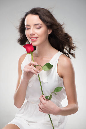 Beautiful woman looks natural and fresh. Her hair fluttering. Her skin is glowing. Her eyes is closed. She holds a rose in handの写真素材