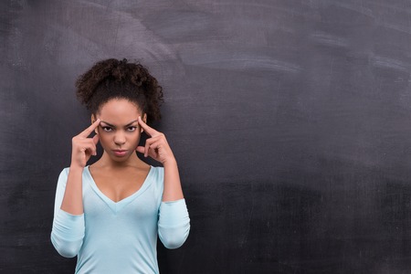 Photo of young pensive afro-american woman on blank chalkboard background. Woman thoughtfully holding point fingers on foreheadの写真素材